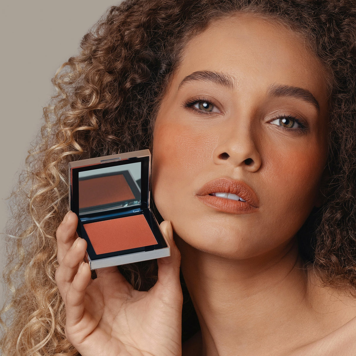 Eine Frau hält den geöffneten Skin Mate Compact von Mesauda Beauty in der Nähe ihres Gesichts und blickt in Kamera.