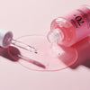 Eine Flasche Peach 70 % Niacinamide Serum mit rosa Flüssigkeit, die mit einer Pipette auf eine hellrosa Oberfläche gekippt wurde.