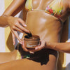 Eine Frau im geblümten Bikini trägt neben einem gelben Handtuch Bali Body Bronze Shimmering Tanning Gel SPF 15 auf ihre strahlende, sonnengeküsste Haut auf.