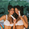 Zwei gebräunte Frauen in weißer Unterwäsche halten lächelnd Face Tan Mist-Flaschen 