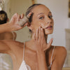 Eine Frau trägt das Tinted Super Serum SPF 30 auf ihre Wange auf, hält die Flasche und blickt in die Kamera