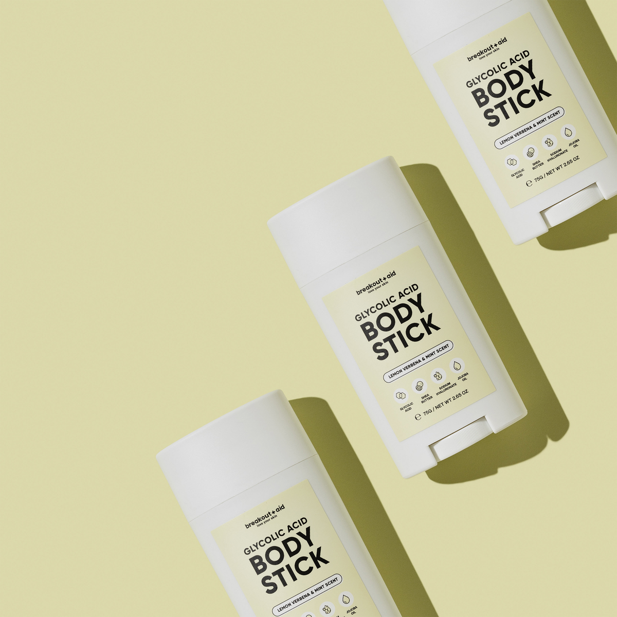 Drei Breakout+Aid Glycolic Acid Body Stick-Hautpflegeprodukte, diagonal auf einem hellgelben Hintergrund angeordnet.