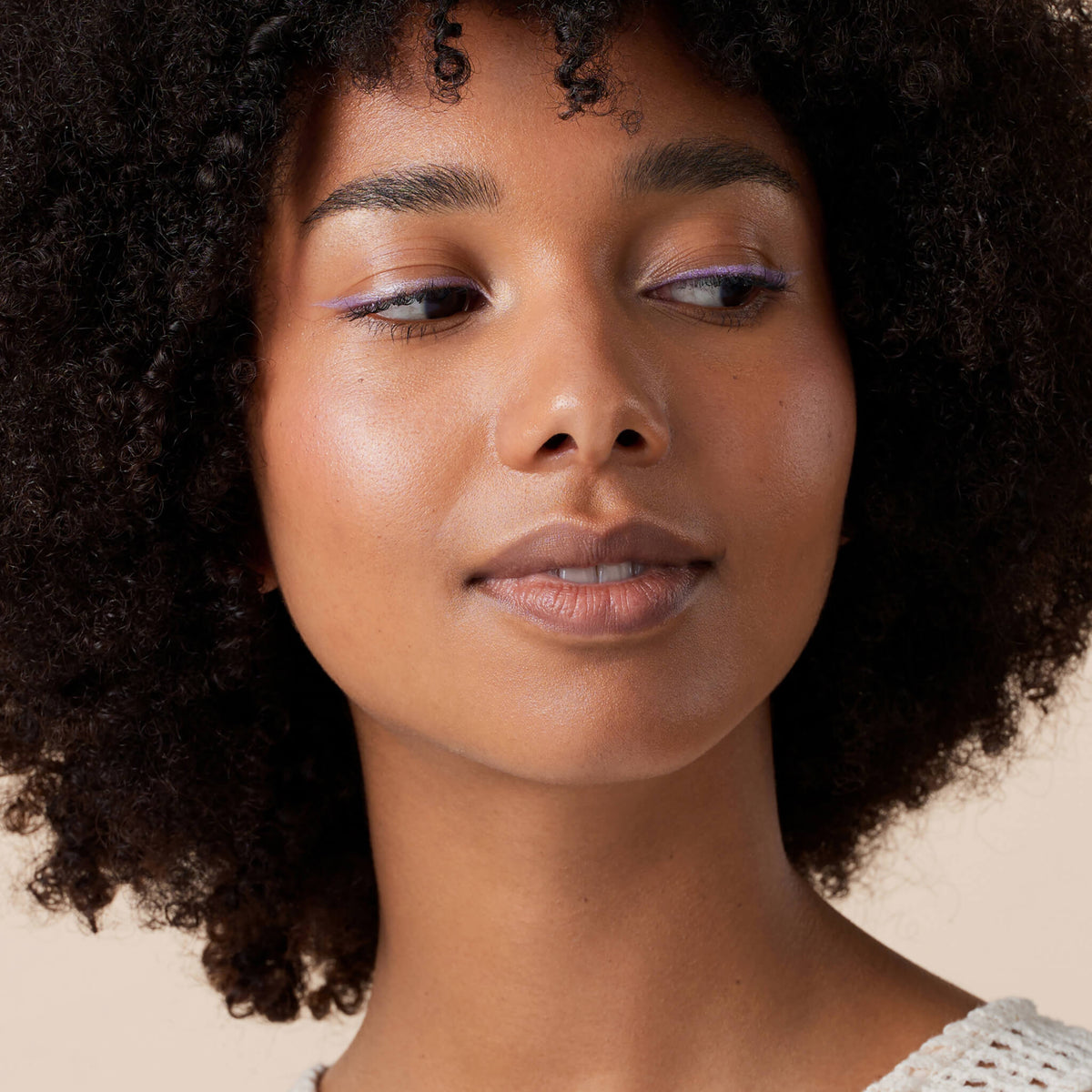 Eine Person mit lockigem Haar trägt den Soft Glam Filter Fluid 040 vor einem neutralen Hintergrund.