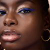 Eine Nahaufnahme zeigt eine Person mit glänzenden Lippen, goldenen Creolen und auffälligem blauen Eyeliner aus der Wonder Woman Eyeshadow Palette.