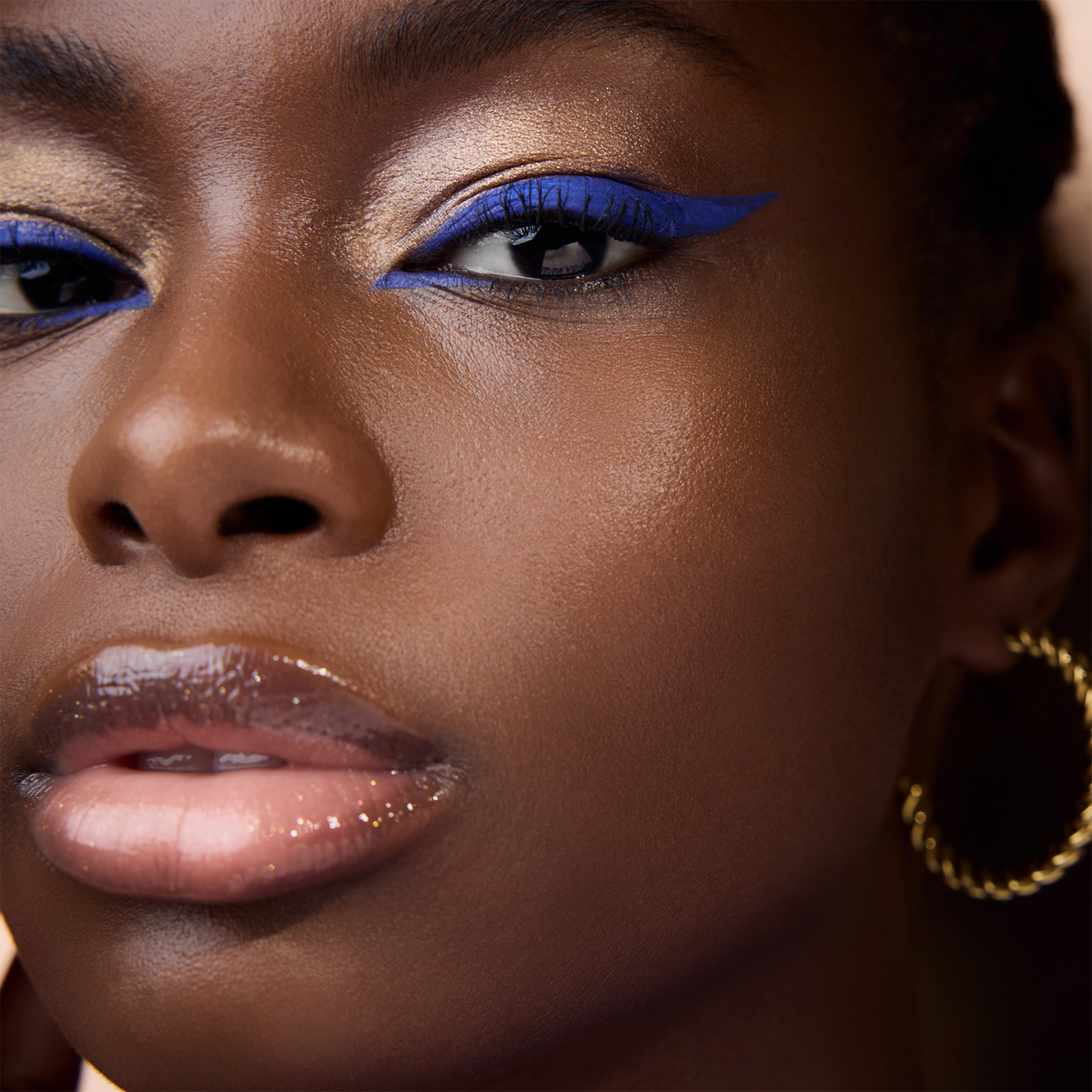 Eine Nahaufnahme zeigt eine Person mit glänzenden Lippen, goldenen Creolen und auffälligem blauen Eyeliner aus der Wonder Woman Eyeshadow Palette.