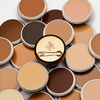 Mehrere geöffnete Tiegel des Yummy Skin Blurring Balm Powder in unterschiedlichen Farben gestapelt; in der Mitte eine geschlossene bronzene Dose mit Löffel.