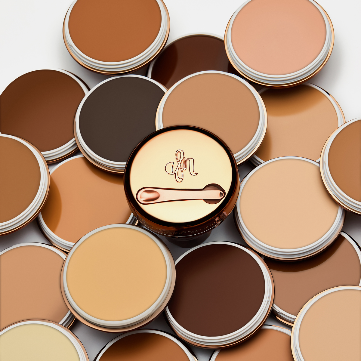 Mehrere geöffnete Tiegel des Yummy Skin Blurring Balm Powder in unterschiedlichen Farben gestapelt; in der Mitte eine geschlossene bronzene Dose mit Löffel.