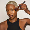 Eine Person mit kurzen blonden Haaren und dunklem Hautton trägt den Yummy Skin Blurring Balm Powder Lowlighter Incognito mit einem Pinsel auf ihre Wange auf.