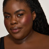 Eine Frau mit dunklem Hautton und schwarzem Haar lächelt und präsentiert den Yummy Skin Blurring Balm Powder Lowlighter Off The Grid. 