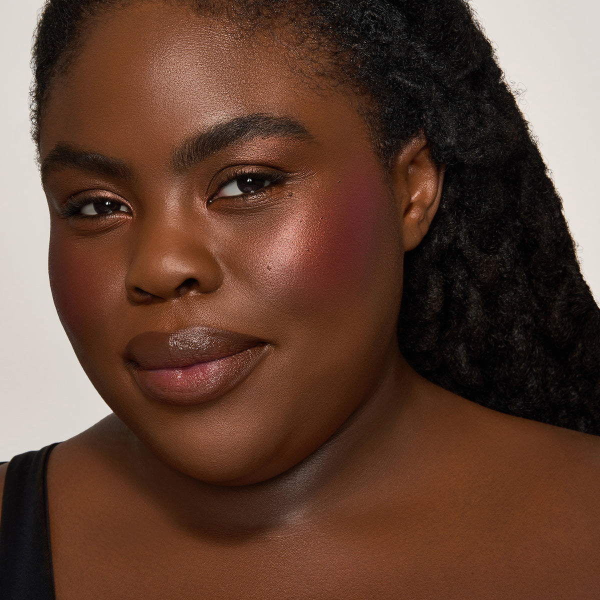 Eine Frau mit dunklem Hautton und schwarzem Haar lächelt und präsentiert den Yummy Skin Blurring Balm Powder Lowlighter Off The Grid. 