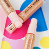Drei Flaschen MAGIC FILTER Glow Booster 40 Tan liegen auf einem bunten geometrischen Hintergrund.