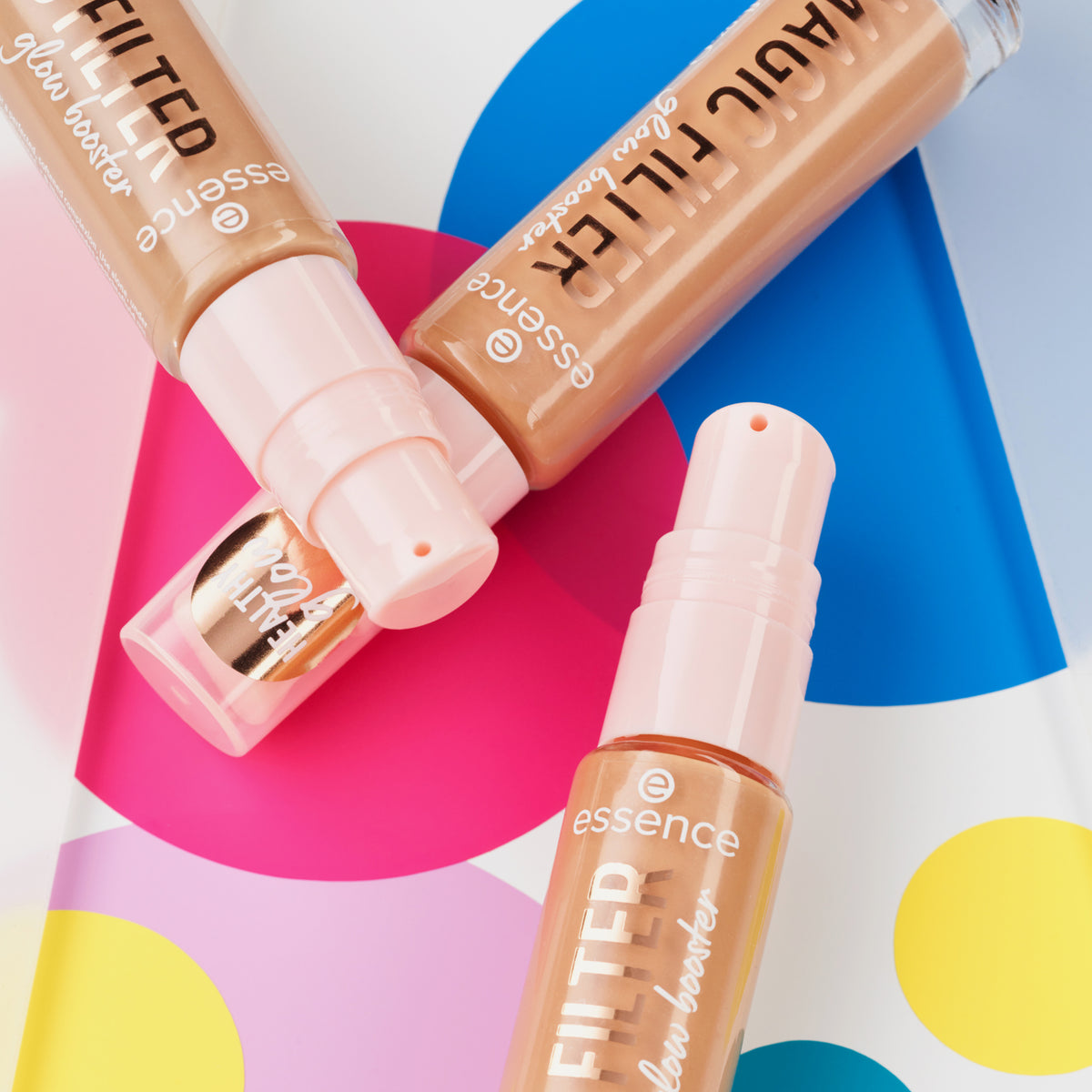 Drei Flaschen MAGIC FILTER Glow Booster 40 Tan liegen auf einem bunten geometrischen Hintergrund.