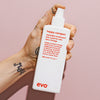 Eine Hand hält eine weiße Flasche Happy Campers Wearable Treatment vor einem hellen Hintergrund.