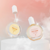 Zwei schwebende Hair-Syrup-Haaröle, Honey Dew und Rose Bloom, vor rosigem Himmel; Glasflaschen glänzen, Etiketten sichtbar.