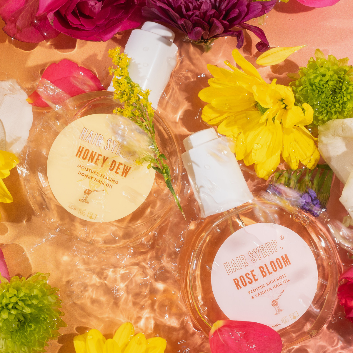 Zwei Hair Syrup Haaröle, Rose Bloom und Honey Dew, liegen im Wasser zwischen bunten Blüten; Etiketten sichtbar, Licht spiegelt sich auf der Oberfläche.
