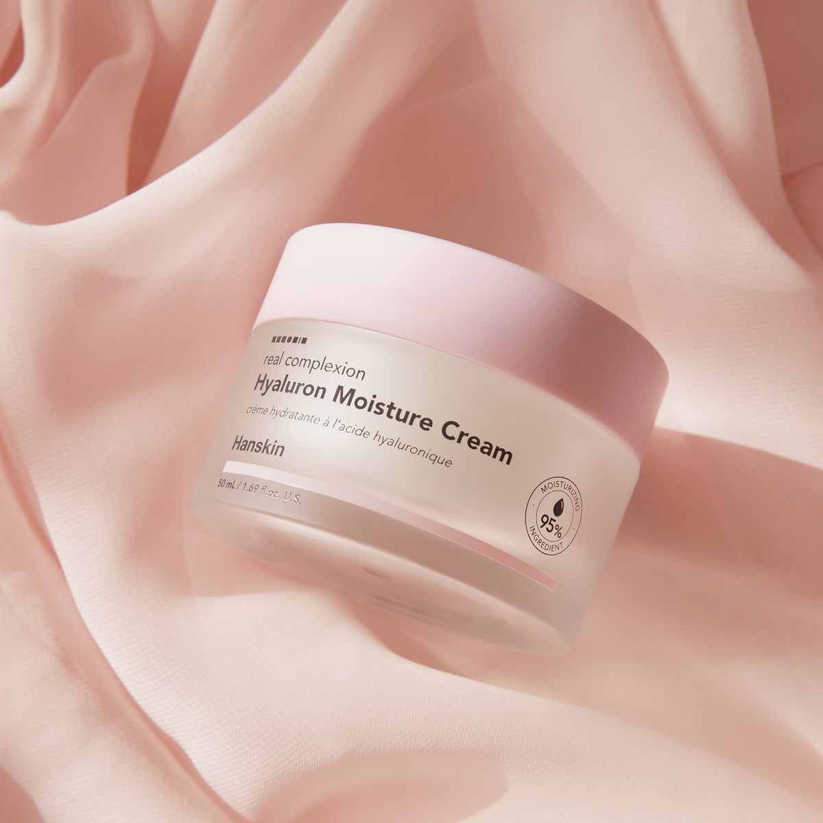 Ein Tiegel Real Complexion Hyaluron Moisture Cream auf einem zartrosa Stoff und wird von sanftem Licht erhellt.