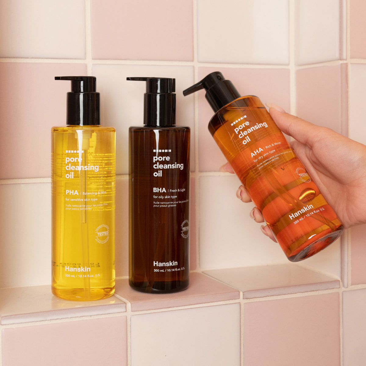 Drei Flaschen Pore Cleansing Oil BHA stehen auf einer gefliesten Oberfläche, eine Flasche wird in einer Hand gehalten.