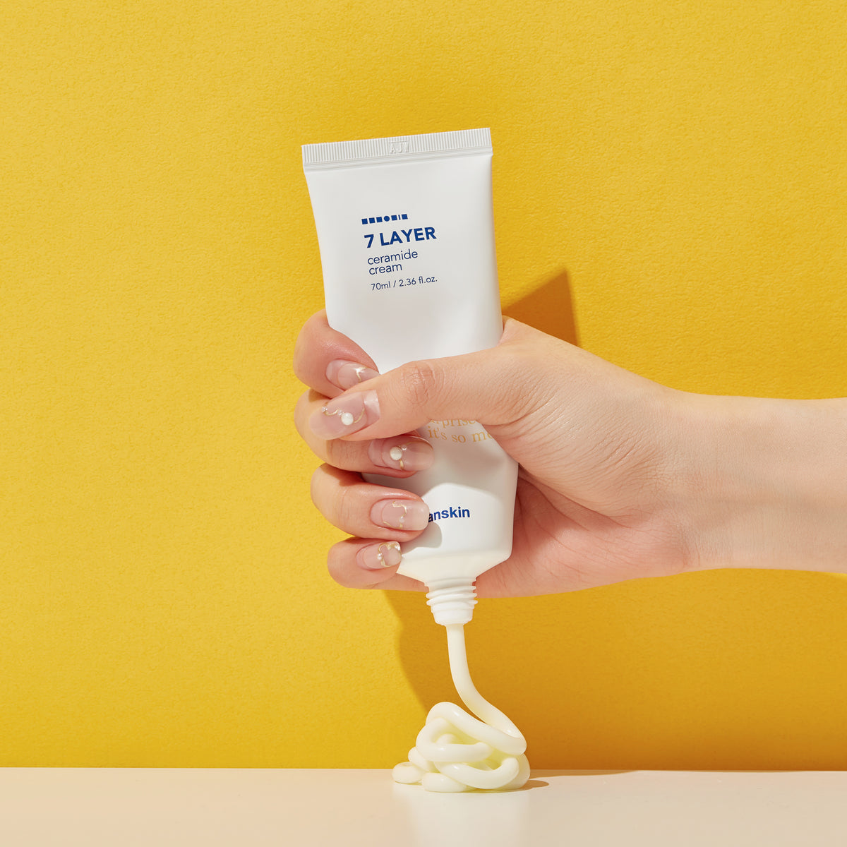 Eine Hand drückt vor einem leuchtend gelben Hintergrund eine Tube Seven Layer Ceramide Cream aus.