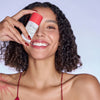 Eine lächelnde Frau mit lockigem Haar hält das Vitamin A Glow Retinol Serum vor ihr Gesicht.
