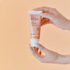 Hände halten Hanskin Vitamin C Glow Mask vor pfirsichfarbenem Hintergrund.