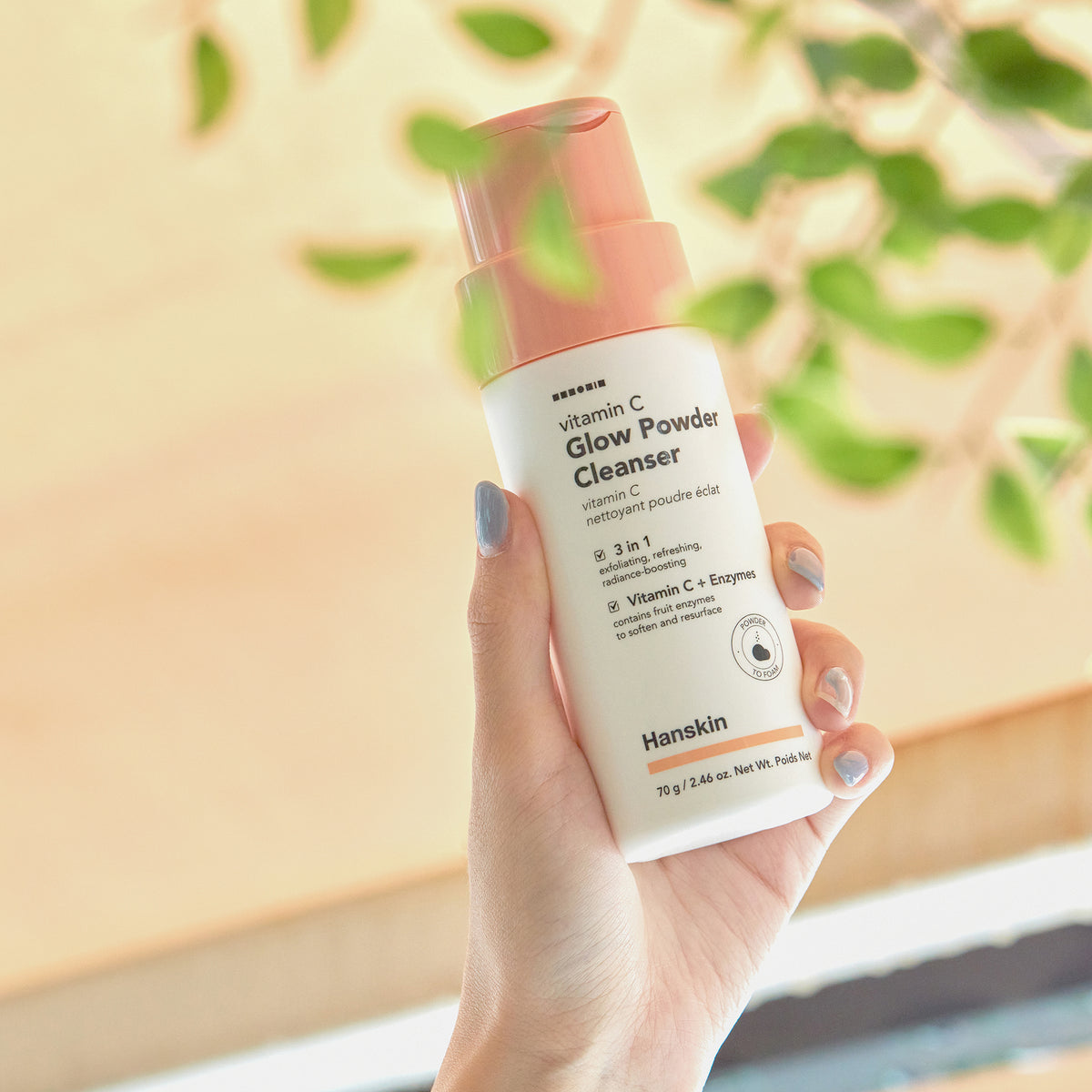 Hand hält eine Flasche Vitamin C Glow Powder Cleanser in der Nähe von grünen Blättern und Sonnenlicht.