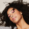 Eine Frau mit lockigem Haar, gestylt mit Living Proof Curl Definer, trägt rosa Lidschatten und goldene Ohrringe, neigt den Kopf und lächelt sanft.