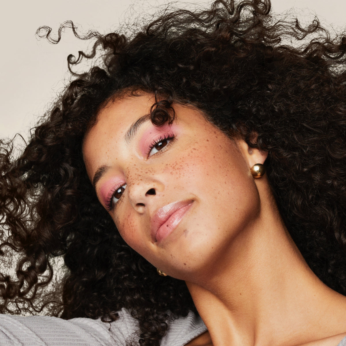Eine Frau mit lockigem Haar, gestylt mit Living Proof Curl Definer, trägt rosa Lidschatten und goldene Ohrringe, neigt den Kopf und lächelt sanft.