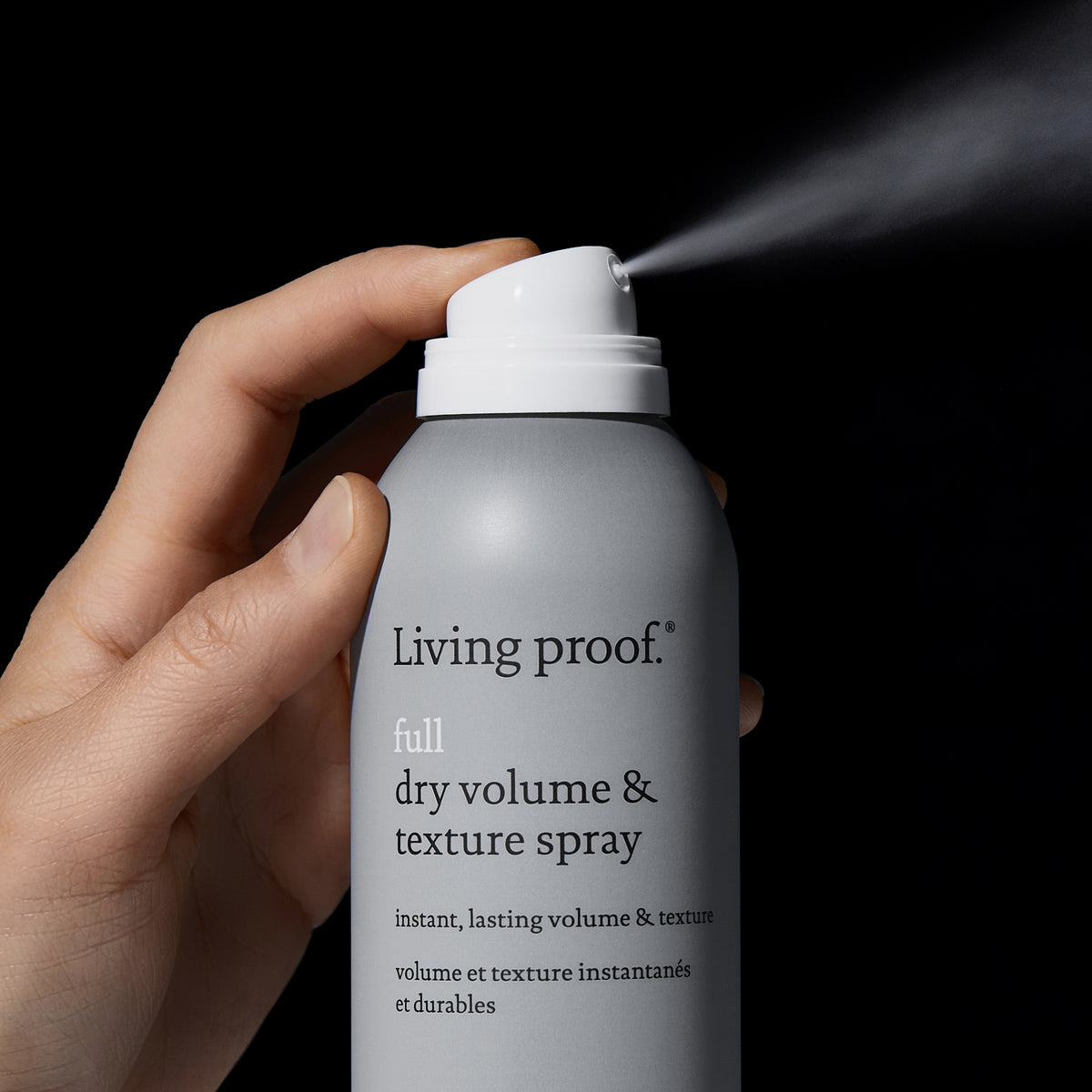 Eine Hand sprüht Living Proof Full Dry Volume & Texture Spray vor einem schwarzen Hintergrund.