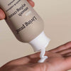 Eine Hand mit offener Handfläche gibt Living Proof No Frizz Smooth Styling Cream aus einer beigen Flasche aus.