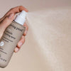 Eine Hand sprüht Living Proof No Frizz Smooth Styling Spray; der Nebel ist vor einem neutralen Hintergrund sichtbar.