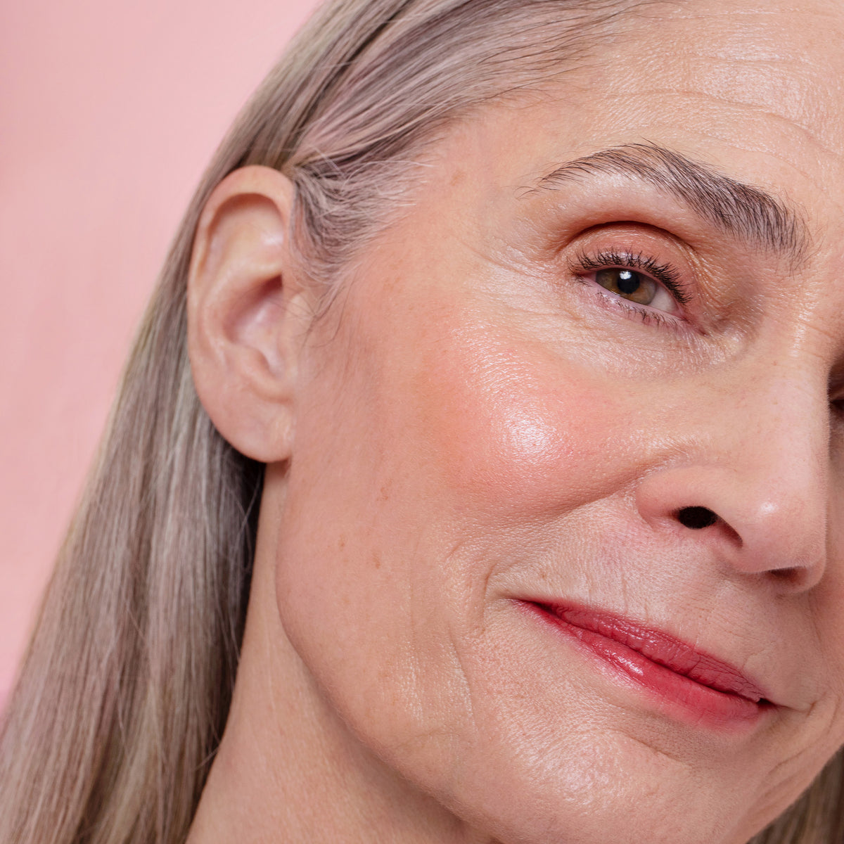 Nahaufnahme einer älteren Frau mit grauem Haar und natürlichem Make-up, die vor einem rosafarbenen Hintergrund sanft lächelt und Lumene INVISIBLE ILLUMINATION Liquid Blush Pink Blossom trägt.