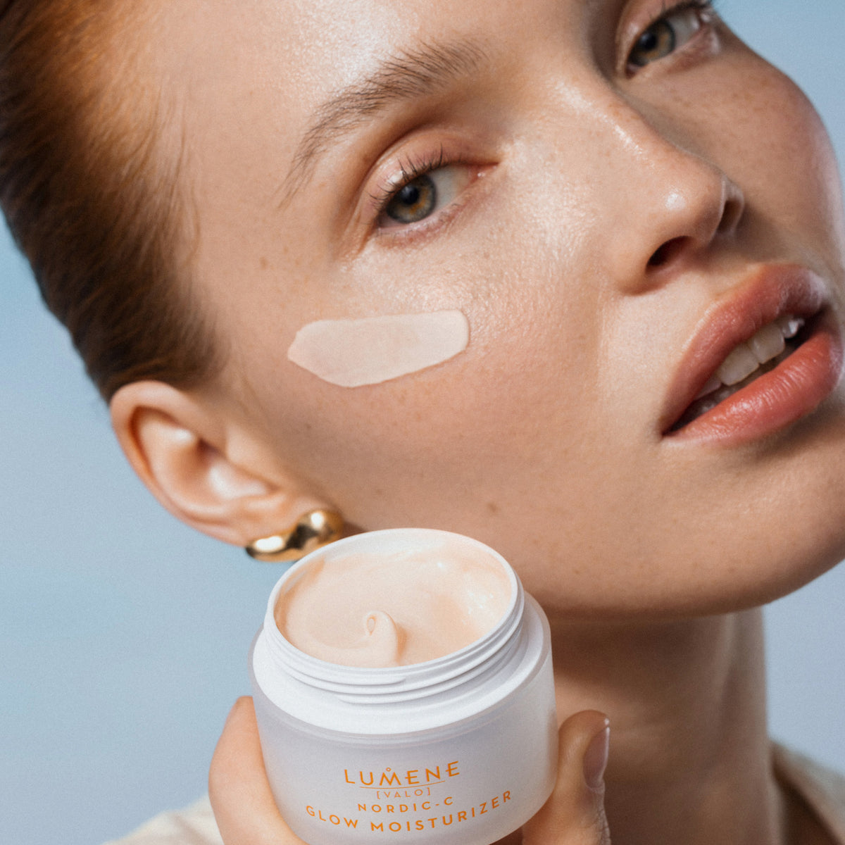 Eine Frau hat einen Cremestrich des Lumene NORDIC-C [VALO] Glow Moisturizer auf der Wange und hält den offenen Tiegel in der Hand.