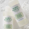 Zwei Flaschen Mario Badescu Glycolic Foaming Cleanser liegen auf einer blubbernden, seifigen Oberfläche.