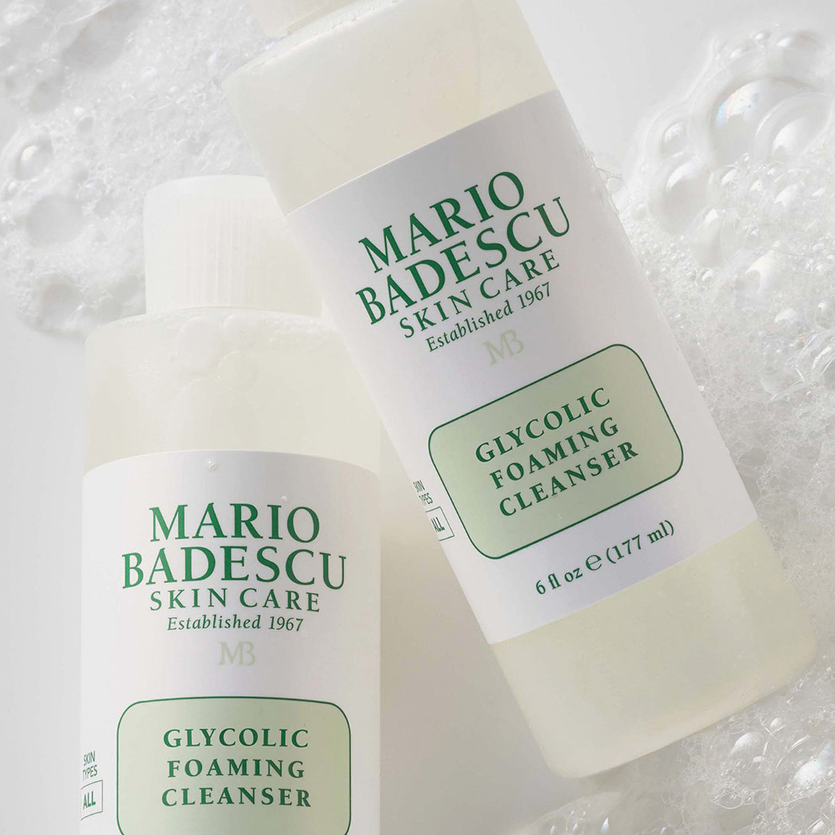 Zwei Flaschen Mario Badescu Glycolic Foaming Cleanser liegen auf einer blubbernden, seifigen Oberfläche.