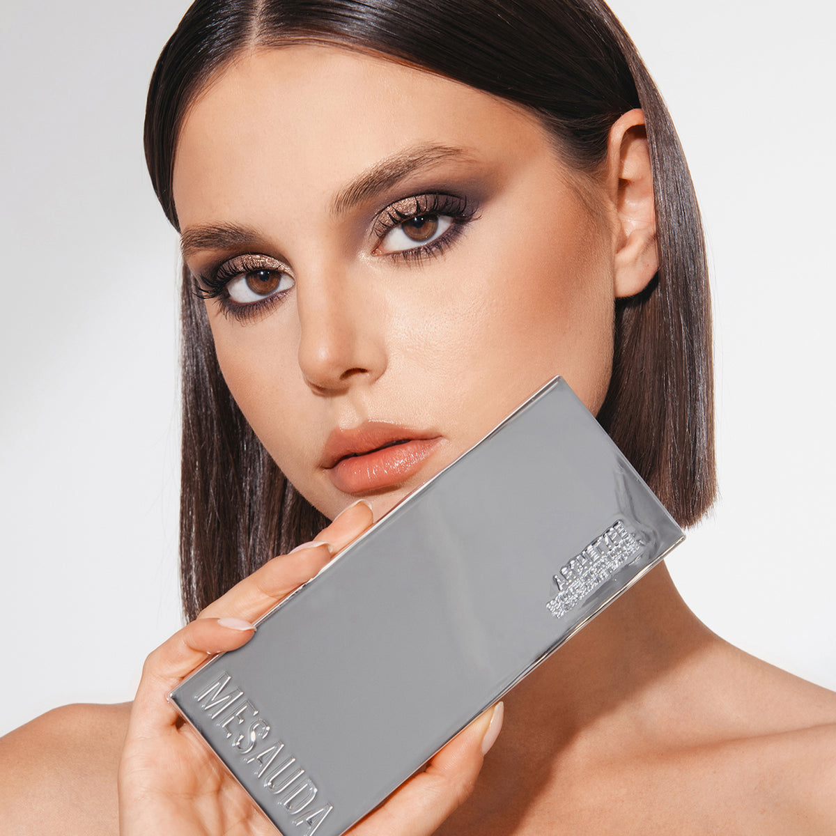 Frau mit glattem braunem Haar hält die silbern glänzende ARCHETYPE Makeup-Palette von Mesauda Beauty an ihr Gesicht.