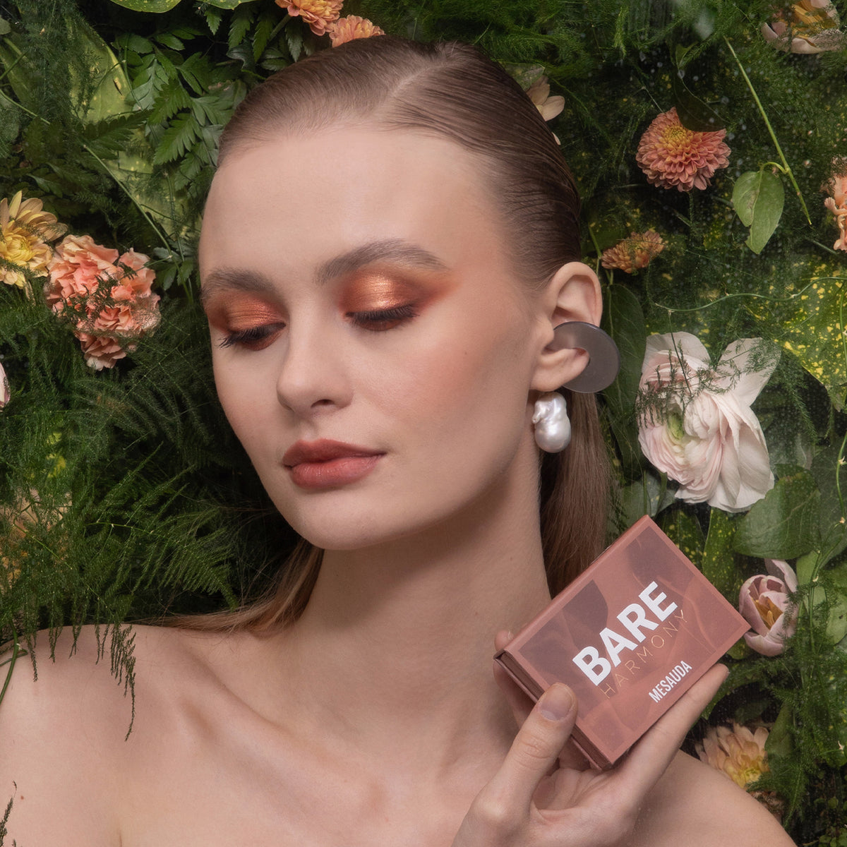 Eine Frau hält die Mesauda Beauty Bare Harmony 3.0 Make-up Box in der Hand, umgeben von Blumen und Grünzeug, die Augen geschlossen.