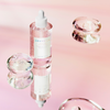 Eine rosafarbene Ampullenflasche mit weißer Pipette steht auf spiegelnder Oberfläche, umgeben von glasklaren Gelkugeln vor rosa Hintergrund.