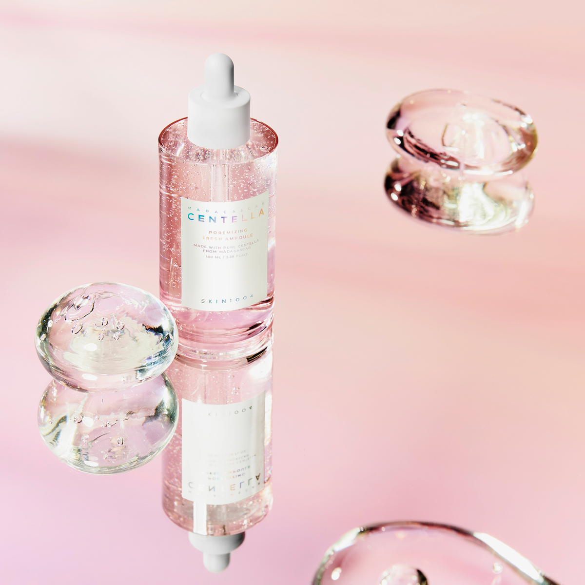 Eine rosafarbene Ampullenflasche mit weißer Pipette steht auf spiegelnder Oberfläche, umgeben von glasklaren Gelkugeln vor rosa Hintergrund.