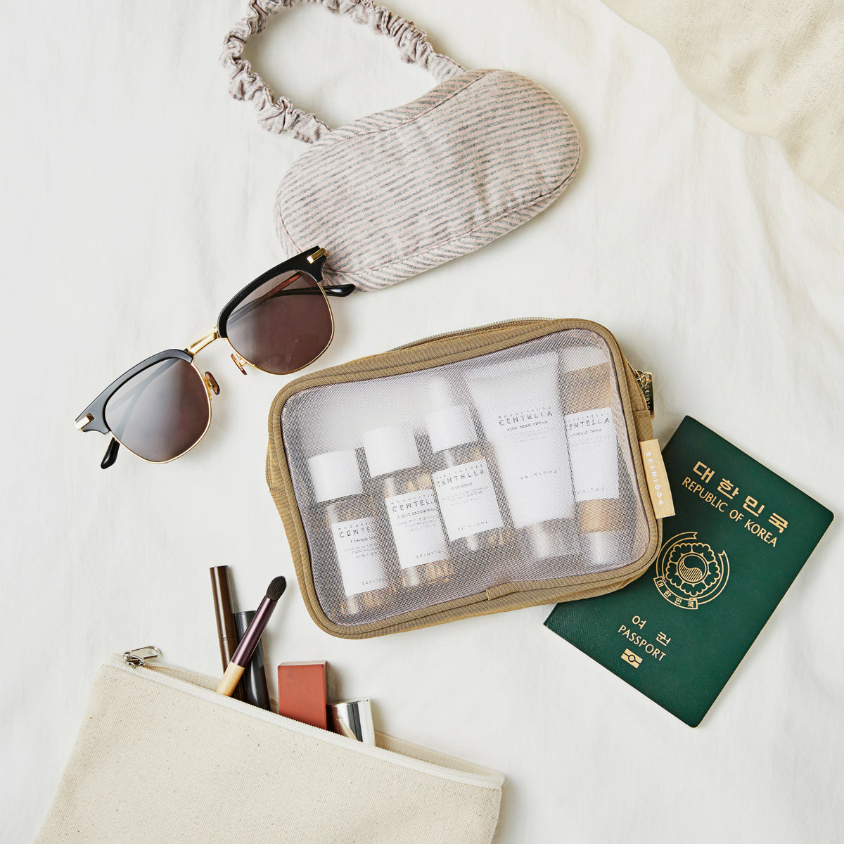 Reiseutensilien auf einem Bett: Sonnenbrille, Augenmaske, koreanischer Reisepass, Kulturbeutel, Kosmetiktasche und das Madagascar Centella Travel Kit.