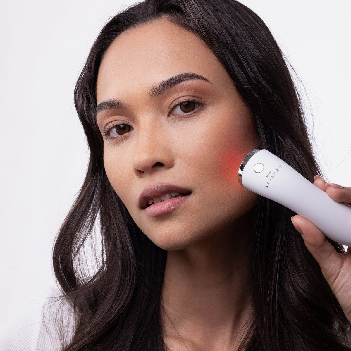 Eine Frau benutzt den STYLPRO Brightspot Anti-Blemish LED-Stab, der ein rotes Licht auf ihre Wange abstrahlt, vor einem schlichten Hintergrund.