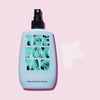 Eine blaue Flasche Hydrating Body Spray mit einem weißen Spritzer daneben; auf rosa Hintergrund.