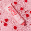 Eine Tube Rapid Warming Balm auf einer rosa Wasseroberfläche, umgeben von schwimmenden Kirschen.