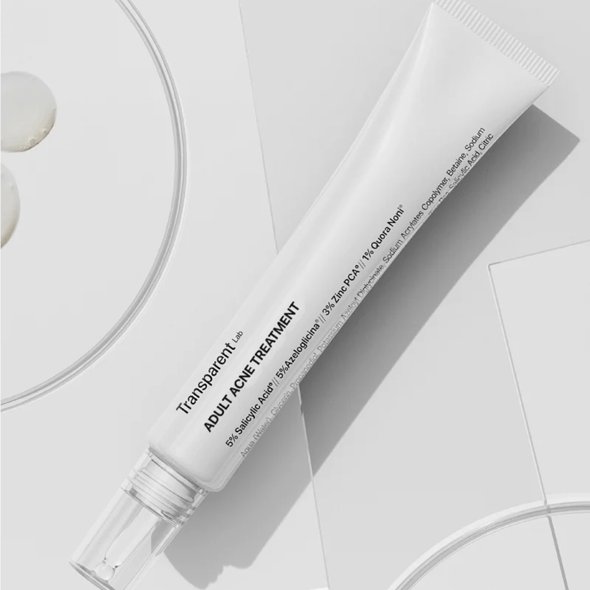 Eine weiße Tube Adult Acne Treatment liegt auf einer weißen Oberfläche mit Schatten und einem Teil einer runden Schale im Blick.