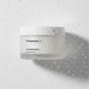 Ein Glas Transparent Lab Cleansing Balm steht auf einer nassen weißen Oberfläche, die mit Wassertropfen bedeckt ist.