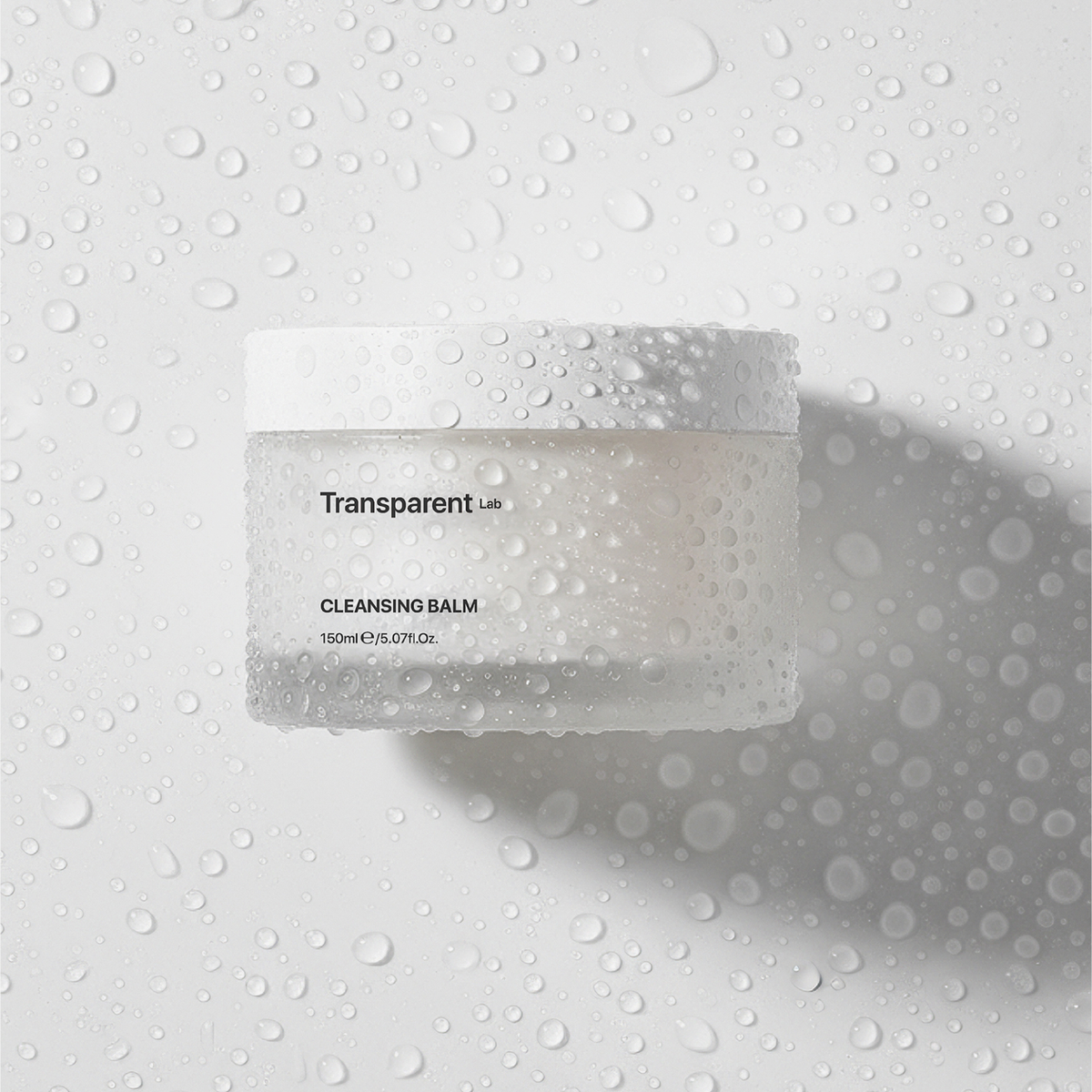 Ein Glas Transparent Lab Cleansing Balm steht auf einer nassen weißen Oberfläche, die mit Wassertropfen bedeckt ist.