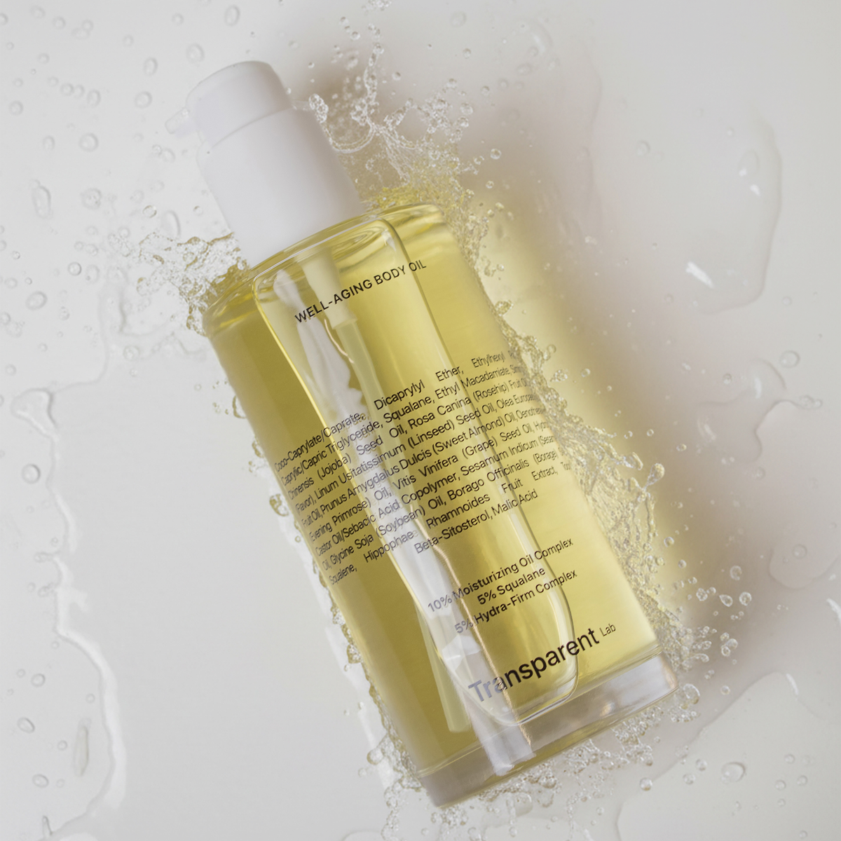 Klare Flasche Well-Aging Body Oil von Transparent Lab mit Wassertropfen auf einer weißen Oberfläche.