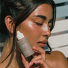 Eine Frau mit strahlender Haut hält eine Flasche Tinted Super Serum SPF 30 in die Kamera und wird vom Sonnenlicht angestrahlt