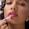 Frau trägt mit einem Applikator ein glänzendes pinkes Lip Oil auf ihre Lippen auf, Nahaufnahme mit Fokus auf Lippen und Produkt.
