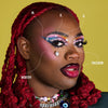 Eine Person mit dunklem Teint mit auffälligem Make-up aus der Love is Love-Palette und roten Zöpfen.