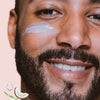 Lächelnder bärtiger Mann mit HUSH Multi-Purpose Moisturiser auf der Wange, neben Symbolen von Kokosnuss, Aloe und einem Wassertropfen.